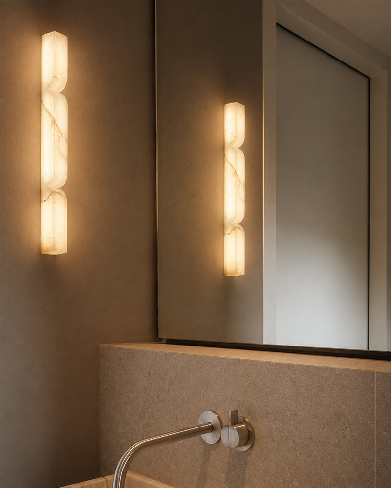 A modern wall-mounted sconce with three vertically stacked frosted rectangular light bars in a warm cream tone, shown installed on a bathroom wall beside a mirror and sink faucet.