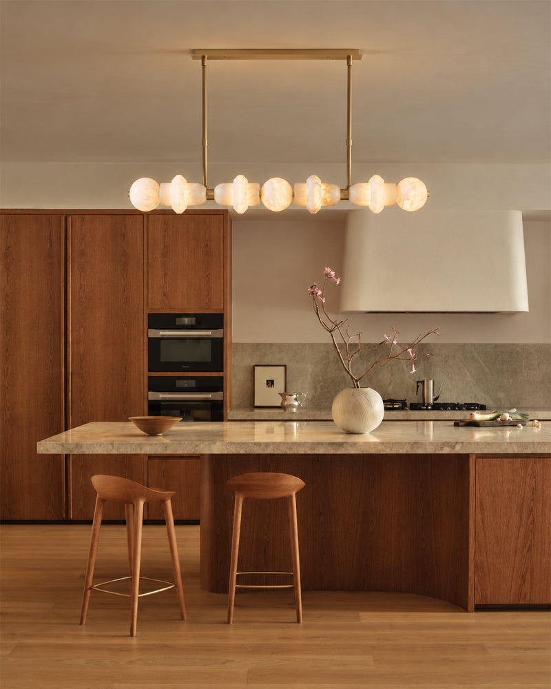 A modern kitchen interior with a brass linear pendant light featuring multiple frosted glass globes, hanging above a marble-topped island with two wooden bar stools.