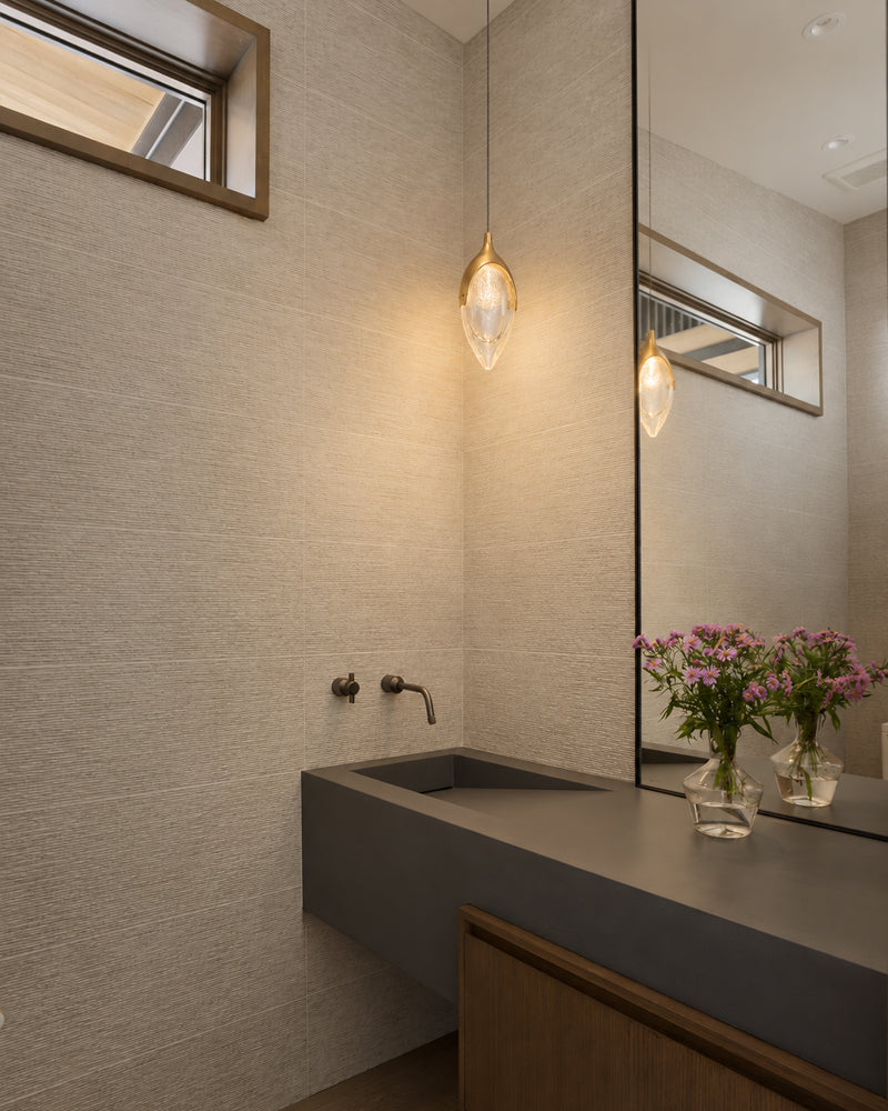 Modern bathroom vanity with a dark gray integrated sink, textured beige walls, a hanging glass pendant light, and a large mirror reflecting the space. Two small glass vases with pink flowers sit on the counter.