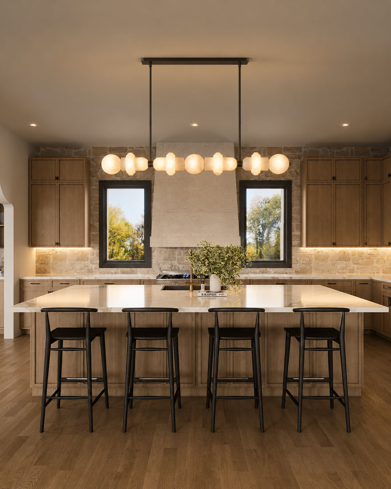 A modern kitchen island with a white stone countertop, light wood base, five black bar stools, and a black linear pendant light with multiple spherical glass shades hanging above. The kitchen has wood cabinets, a stone backsplash, and two windows.