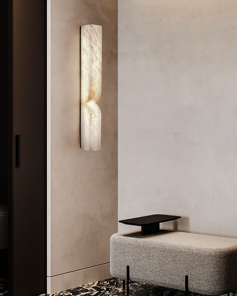 Sleek alabaster wall sconce illuminating a minimalist entryway with gray bench and black table.