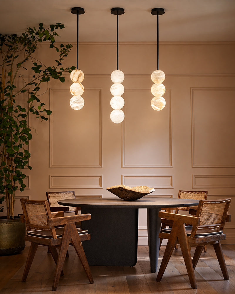 Three modern pendant light fixtures with stacked spherical glass shades hang above a round dining table with wooden chairs in a warm, elegant room.