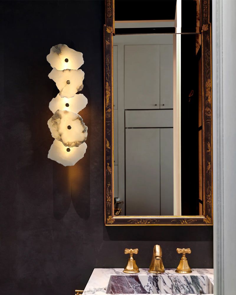 A polished brass wall sconce with a white alabaster shade, featuring a multi-head design with an irregular layout, mounted on a wall above a bathroom sink and mirror.