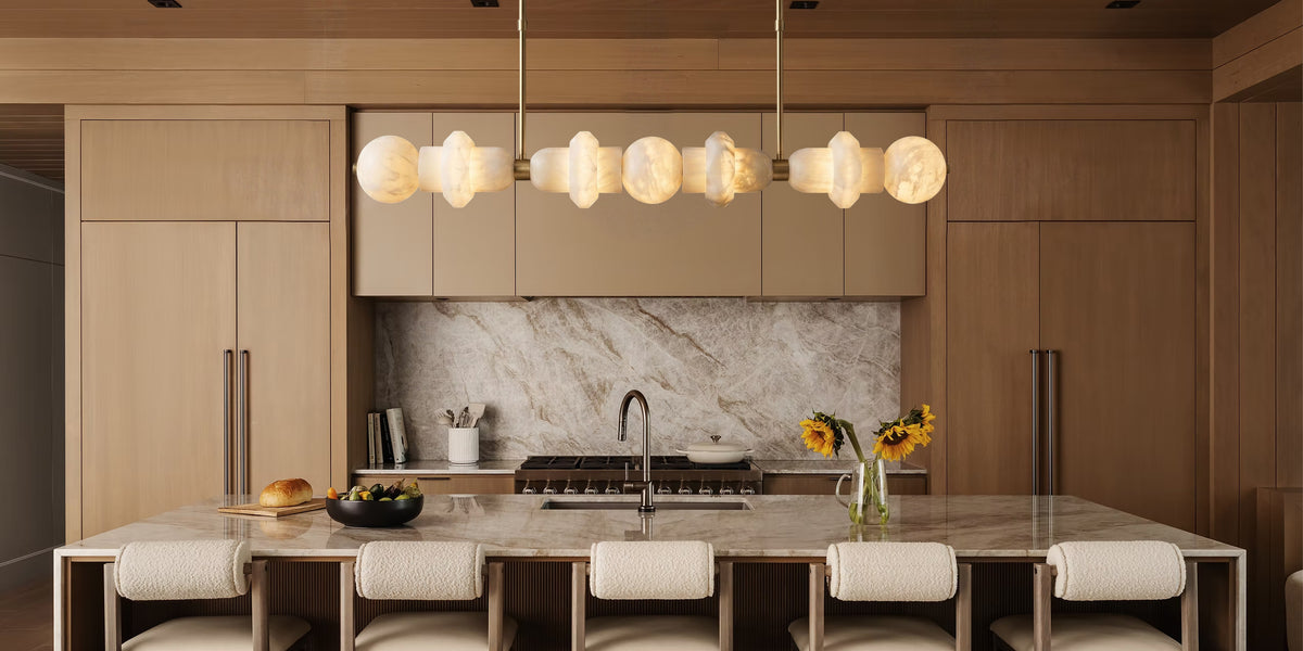 A modern kitchen interior with warm wood cabinetry, a marble backsplash, a marble island countertop, and a row of upholstered bar stools beneath a multi-light pendant fixture.