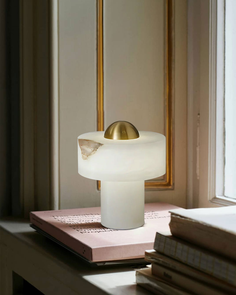 Stylish alabaster table lamp with brass detail on a stack of books by a window.