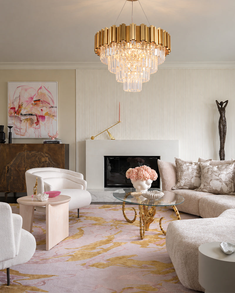 A luxury modern living room featuring a prominent gold and crystal chandelier hanging above a glass-top coffee table. The room is styled with a beige sectional sofa, white accent chairs, a pink and gold patterned rug, and elegant wall art.