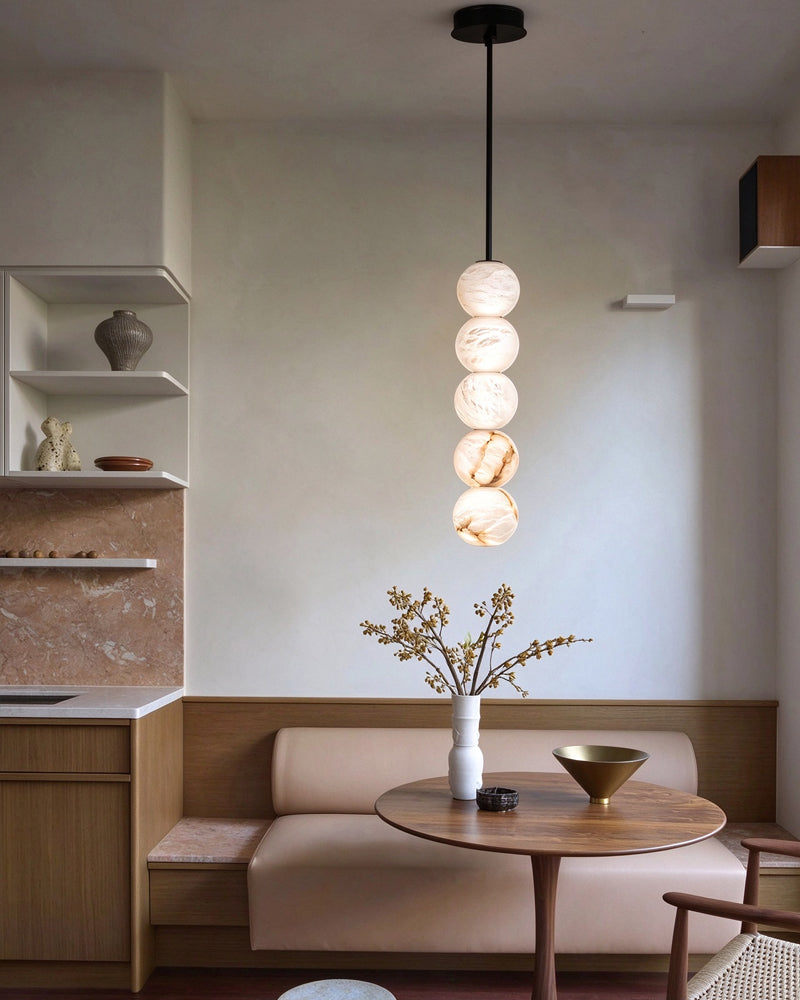 A modern multi-light pendant fixture with stacked spherical glass shades hangs over a round wooden table in a minimalist dining or living space with a beige upholstered bench and neutral walls.
