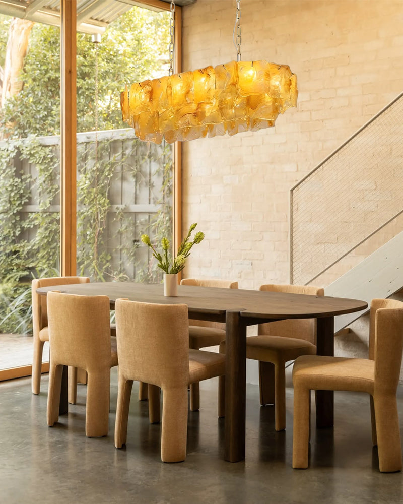 A modern dining room scene with a dark wood oval table and six upholstered dining chairs in a warm beige tone, under a glowing amber glass chandelier. Large windows reveal greenery outside, and a staircase is visible against a light brick wall.