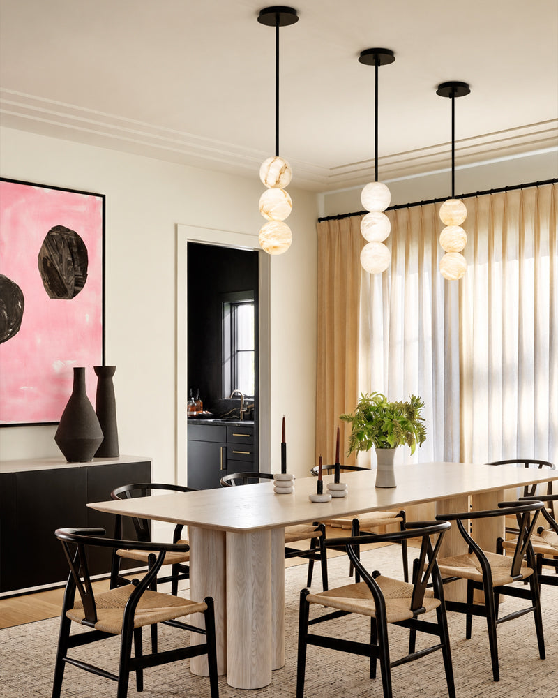 A modern dining room featuring a light wood dining table with woven-back chairs, three black pendant lights with stacked spherical glass shades, beige curtains, and a pink abstract wall art piece.