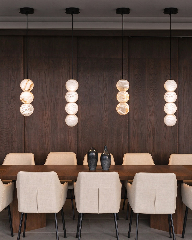 A modern dining room setup featuring a long wooden table surrounded by beige upholstered chairs, with four hanging pendant lights composed of stacked oval glass shades glowing against a dark wood-paneled wall.