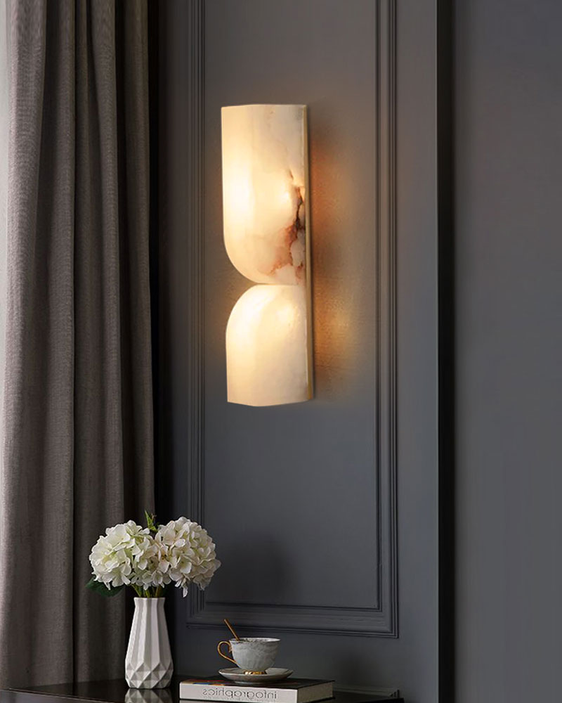 Curved alabaster wall sconce providing warm light on dark wall beside white flower vase.